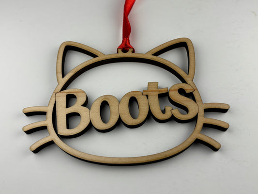 Wooden ornament shaped like a cat's head with 'Boots' engraved, hanging on a red string against a white background.